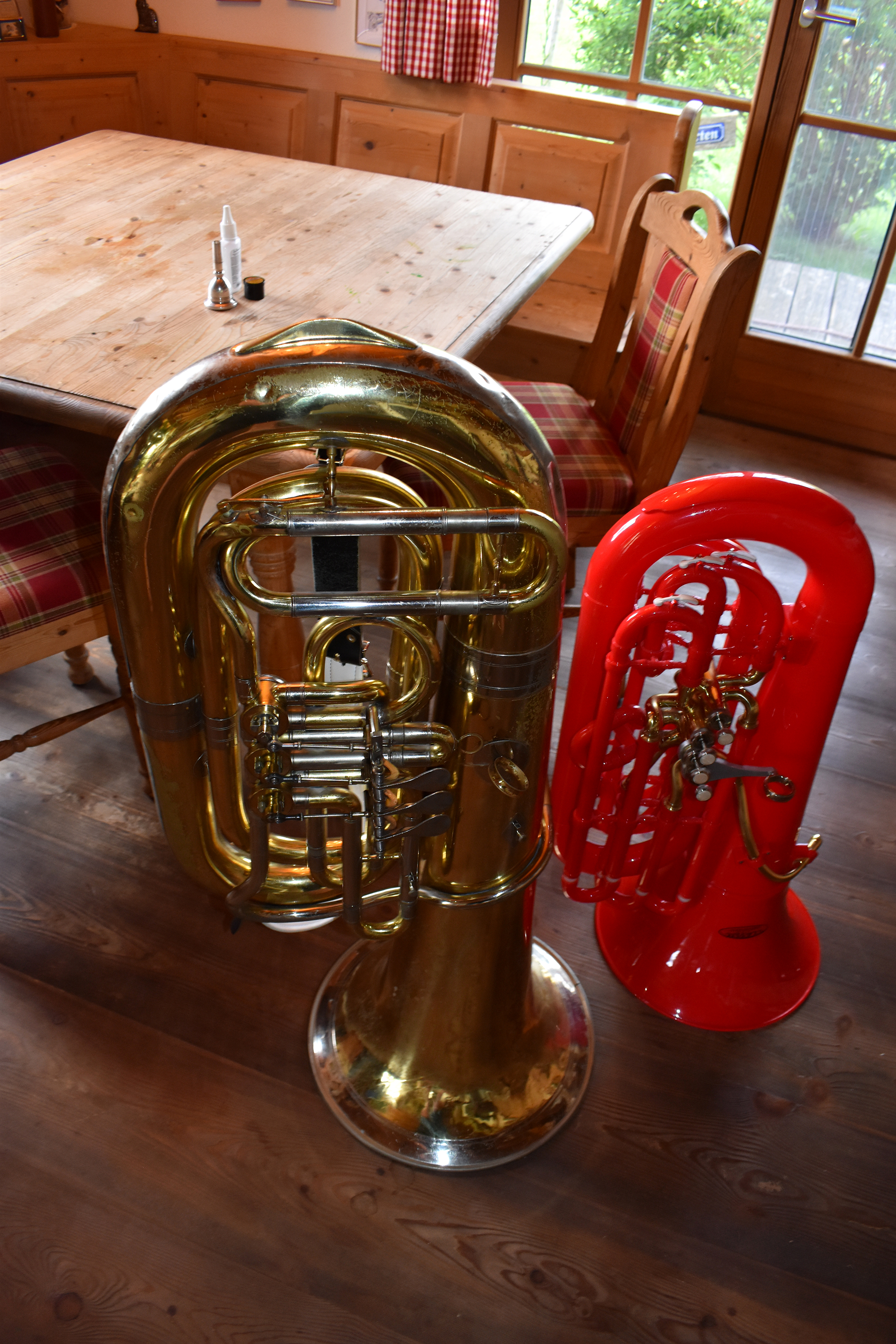 Optischer Vergleich einer normalen Tuba aus Messing mit der MardiBrass-Tuba aus rotem Kunststoff. Die beiden Tuben stehen nebeneinander in einer bayerischen Stube auf einem Holzfußboden. Man sieht deutlich, dass die Plastiktuba kompakter ist als das Modell aus Messing.